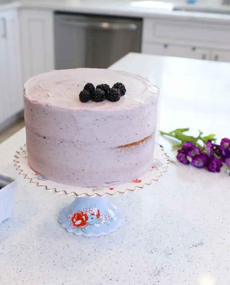 A tall three-layer Blackberry Lemon Cake decorated with fresh berries on a vintage cake stand.