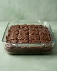 A square chocolate zucchini cake with chocolate chips and a glossy glaze inside a baking pan.