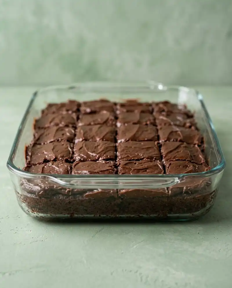 A square chocolate zucchini cake with chocolate chips and a glossy glaze inside a baking pan.