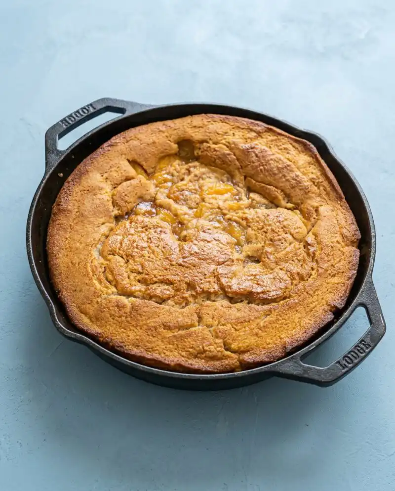 A golden brown Classic Peach Cobbler Dump Cake bubbling in a glass baking dish on a wooden table.
