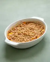 A golden brown Peach Crisp bubbling in a white ceramic baking dish on a wooden table.