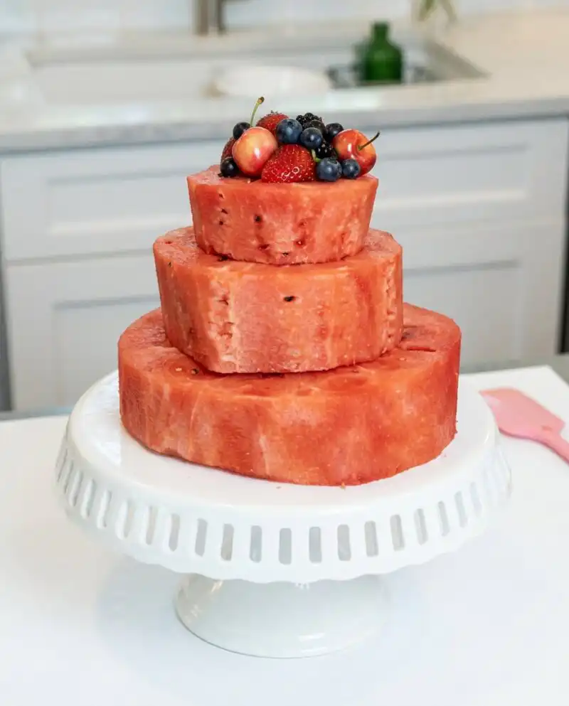 A three tiered Fresh Watermelon Layer Cake decorated with berries and mint on a white stand.