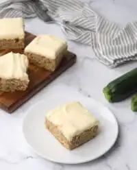 A square slice of Zucchini Bars topped with thick cream cheese frosting on a white plate.