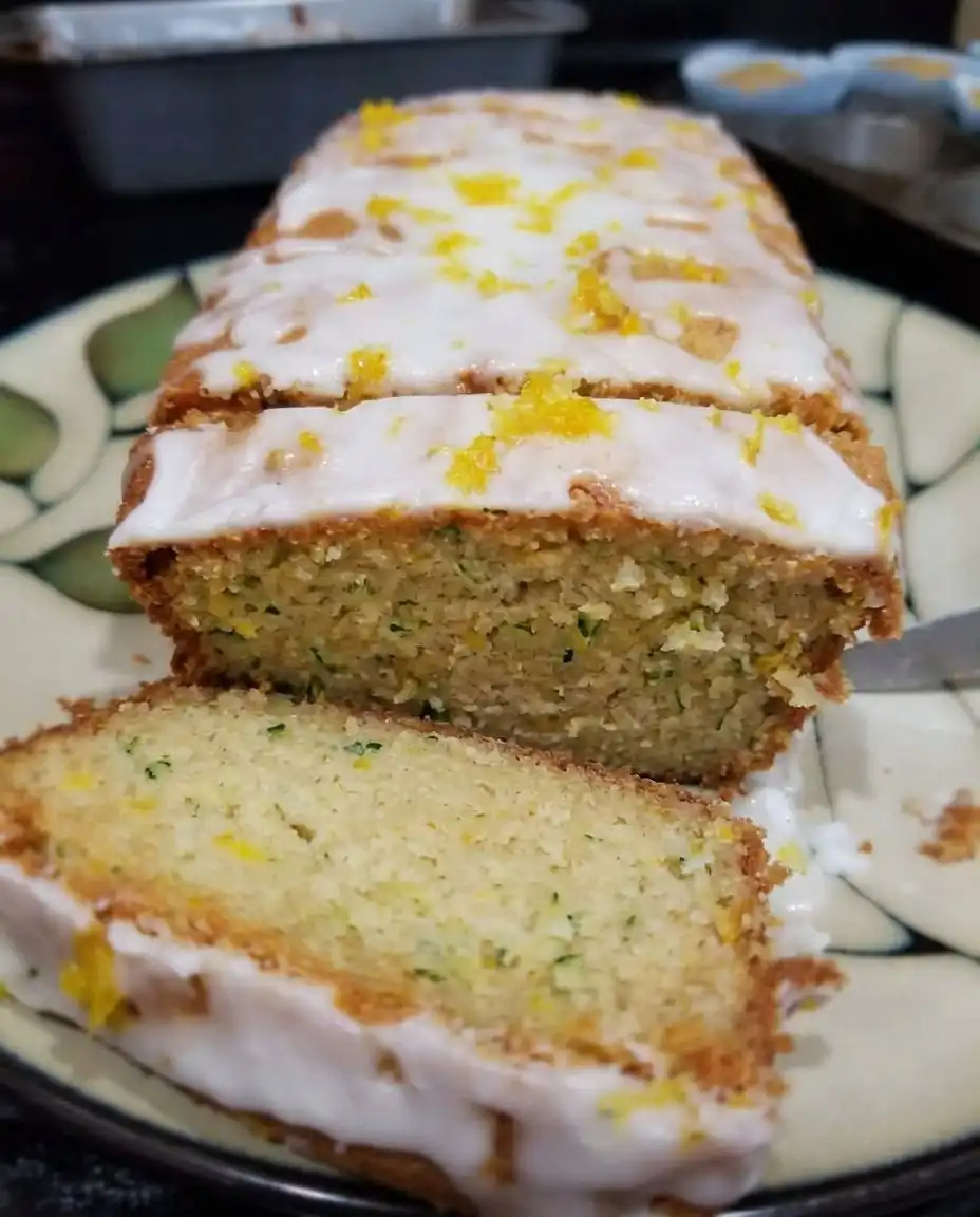 A hand drizzling white lemon glaze over a golden zucchini bread loaf