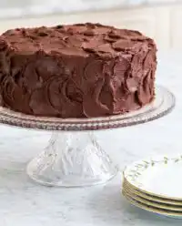 A rustic Devil's Food Layer Cake sitting on a dark wooden cutting board in a farmhouse kitchen.