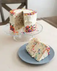 A rustic Funfetti Birthday Layer Cake with white frosting and colorful sprinkles on a wooden board.