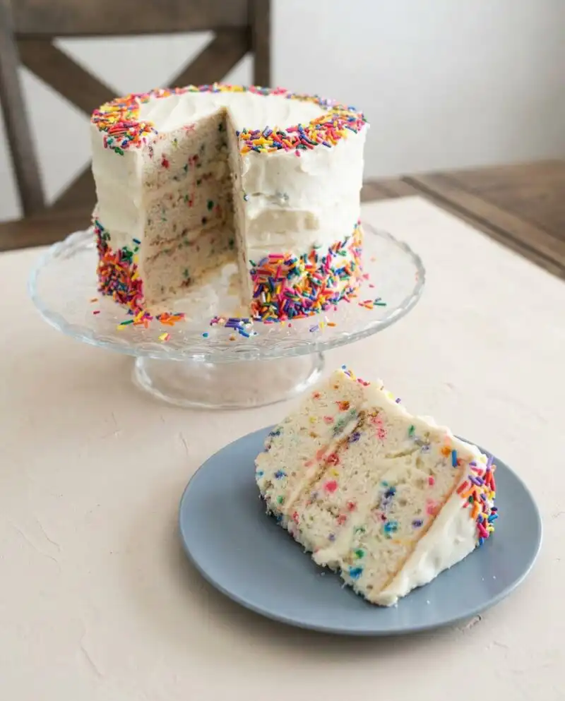 A rustic Funfetti Birthday Layer Cake with white frosting and colorful sprinkles on a wooden board.