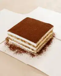 Slices of Classic Tiramisu Layer Cake dusted with cocoa powder on a vintage cake stand.