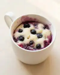 An Easy Blueberry Mug Cake sits in a white ceramic mug on a weathered wooden board.