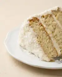 A gorgeous Three-Layer Coconut Cream Cake coated in shredded coconut on a white plate