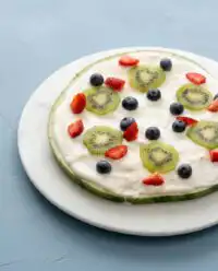 A rustic watermelon pizza cake topped with fresh strawberries and sweet berries on a wooden farmhouse table.